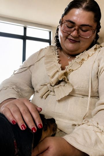 Deb seated in soft window light with a dog curled into her lap, one hand resting gently on the dog&rsquo;s back